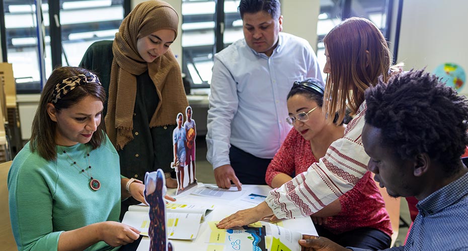 Sechs Menschen sehen das Material des Workshopkoffers durch, das auf einem Tisch ausgebreitet ist.